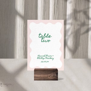 Puede incluir: Una tarjeta de número de mesa rosa con un borde blanco festoneado. La tarjeta dice "mesa dos" en escritura cursiva verde. Debajo del número de mesa, dice "Hannah Prior & Phillip Mackay 25.01.24" en escritura cursiva verde. La tarjeta está sentada en un soporte de madera.