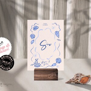 May include: A light pink table number card with a blue seashell design and the number "Six" written in cursive. The card is on a wooden stand.
