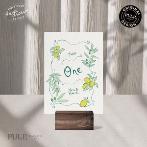 Puede incluir: Una tarjeta de mesa blanca con ilustraciones botánicas verdes de limones y hojas. La tarjeta dice "Table One" y "Maria & Roberto". Está sostenida por un bloque de madera oscura. También se ve el texto "Hand Drawn by Pulp" y "Original Pulp Design".