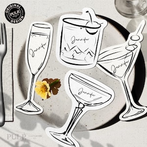 May include: Black and white line art drink-themed place cards with the name "Jennifer" on each. The cards depict a champagne flute, cocktail, martini, and coupe glass. A yellow flower and a fork are also visible.