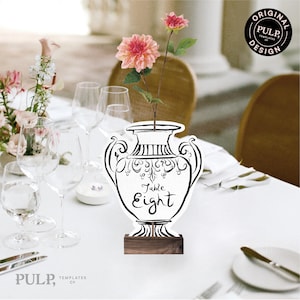 May include: A white table number card in the shape of a vase, with the words "Table Eight" in black script. The card is decorated with black ornate designs and sits on a wooden base. A pink flower is placed in the vase. The image also includes wine glasses and a white tablecloth.