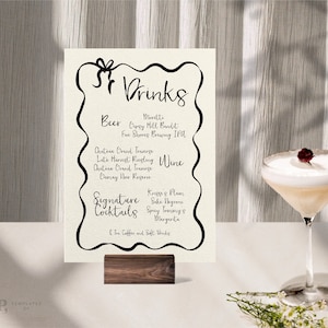 Könnte beinhalten: Ein weißes Schild mit schwarzem Text, auf dem die verfügbaren Getränke aufgelistet sind, darunter Bier, Wein und Signature-Cocktails. Das Schild steht auf einem Holzständer und im Hintergrund ist ein Glas mit einem weißen Cocktail mit einer roten Beere zu sehen.