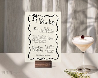 Könnte beinhalten: Ein weißes Schild mit schwarzem Text, auf dem die verfügbaren Getränke aufgelistet sind, darunter Bier, Wein und Signature-Cocktails. Das Schild steht auf einem Holzständer und im Hintergrund ist ein Glas mit einem weißen Cocktail mit einer roten Beere zu sehen.