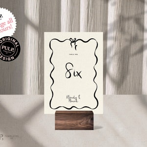 Könnte beinhalten: Eine schwarz-weiße Tischkarten mit einem dekorativen Rand und der Nummer "Six" in Kursivschrift. Die Karte enthält auch die Namen "Wendy & Henrik" in Kursivschrift. Die Karte steht auf einem Holzständer.
