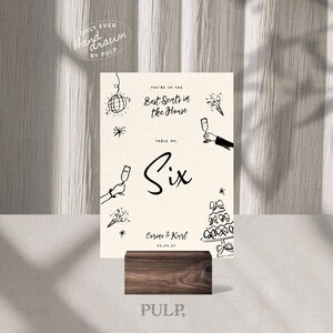 Könnte beinhalten: Eine cremefarbene Tischnummerkarte mit schwarzen Illustrationen und Text, einschließlich des Wortes "Six" in einer großen Schriftart. Die Karte wird von einem dunklen Holzblock gehalten. Der Text "You're in the Best Seats in the House" ist sichtbar.