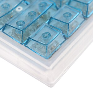 Op de afbeelding: Een close-up van een wit toetsenbord met blauwe transparante toetsen. Het toetsenbord is gemaakt van plastic en heeft een wit frame.