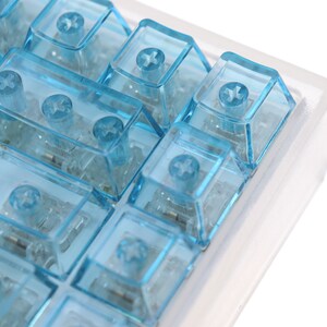 Op de afbeelding: Een close-up van een set blauwe mechanische toetsenbordtoetsen in een doorzichtige plastic behuizing. De toetsen zijn transparant en hebben een kruisvormig ontwerp aan de bovenkant.