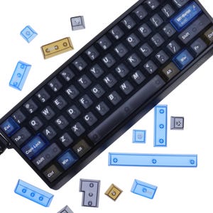 Puede incluir: Un teclado mecánico negro con teclas blancas y una variedad de teclas de repuesto de colores dispersas. El teclado tiene un diseño compacto con una mezcla de teclas estándar y personalizadas.