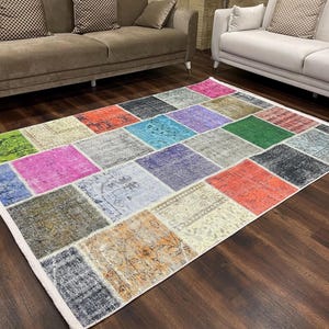 May include: A colourful patchwork rug with a white background. The rug is made up of squares of different colours and patterns, including pink, green, blue, and grey. The rug is laid out on a wooden floor.