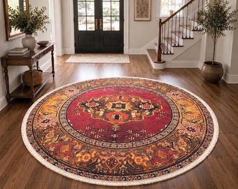 Puede incluir: Una alfombra redonda con un medallón central de color rojo intenso con intrincados patrones geométricos y florales en tonos naranja, negro y crema. La alfombra tiene un borde decorativo ancho y un fleco blanco, colocada sobre un suelo de madera.