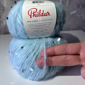 Pu&ograve; includere: Un gomitolo di filato azzurro chiaro con paillettes argentate. Il filato &egrave; etichettato "Phildar Phil Gloria Paillette et Chaleur" e elenca la composizione del filato come 42% poliammide, 21% acrilico, 20% alpaca, 10% poliestere, 7% lana. Il gomitolo pesa 50g (1,76 oz).