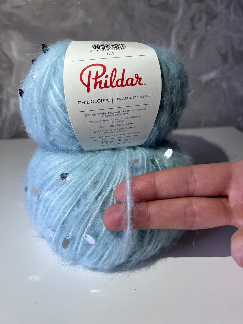 Pu&ograve; includere: Un gomitolo di filato azzurro chiaro con paillettes argentate. Il filato &egrave; etichettato "Phildar Phil Gloria Paillette et Chaleur" e elenca la composizione del filato come 42% poliammide, 21% acrilico, 20% alpaca, 10% poliestere, 7% lana. Il gomitolo pesa 50g (1,76 oz).