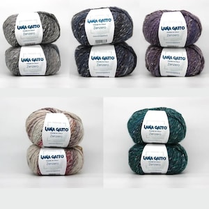 May include: Six skeins of Lana Gatto Zenzero yarn in various colors, including gray, blue, purple, and green. Each skein has a speckled texture and a label with the brand name and product details. Ideal for knitting and crochet projects.