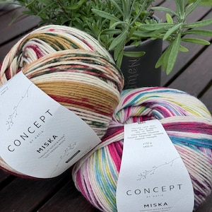 Pode incluir: Duas bolas de fio, uma com um esquema de cores vermelho, branco e verde e a outra com um esquema de cores arco-íris. Ambas as bolas de fio são etiquetadas com "Concept by Katia Miska" e "Premium Socks & More" em texto branco em um rótulo branco.