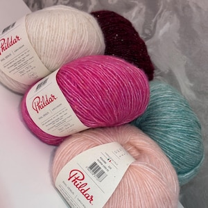 Può includere: Una collezione di gomitoli di lana colorati. L'immagine presenta gomitoli di filato nei toni del bianco, rosa, verde acqua e bordeaux. Il filato è etichettato "Phil Venus" e sembra essere un materiale morbido e strutturato, adatto per progetti di maglieria o uncinetto.