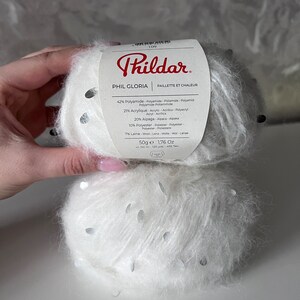 Pu&ograve; includere: Un gomitolo di filato bianco con paillettes argentate, etichettato "Phil Gloria" con una descrizione della composizione del filato: 42% Poliammide, 27% Acrilico, 20% Alpaca, 10% Poliestere, 7% Lana. L'etichetta riporta anche "Paillete et Chaleur" e "50g e 1,76 Oz".