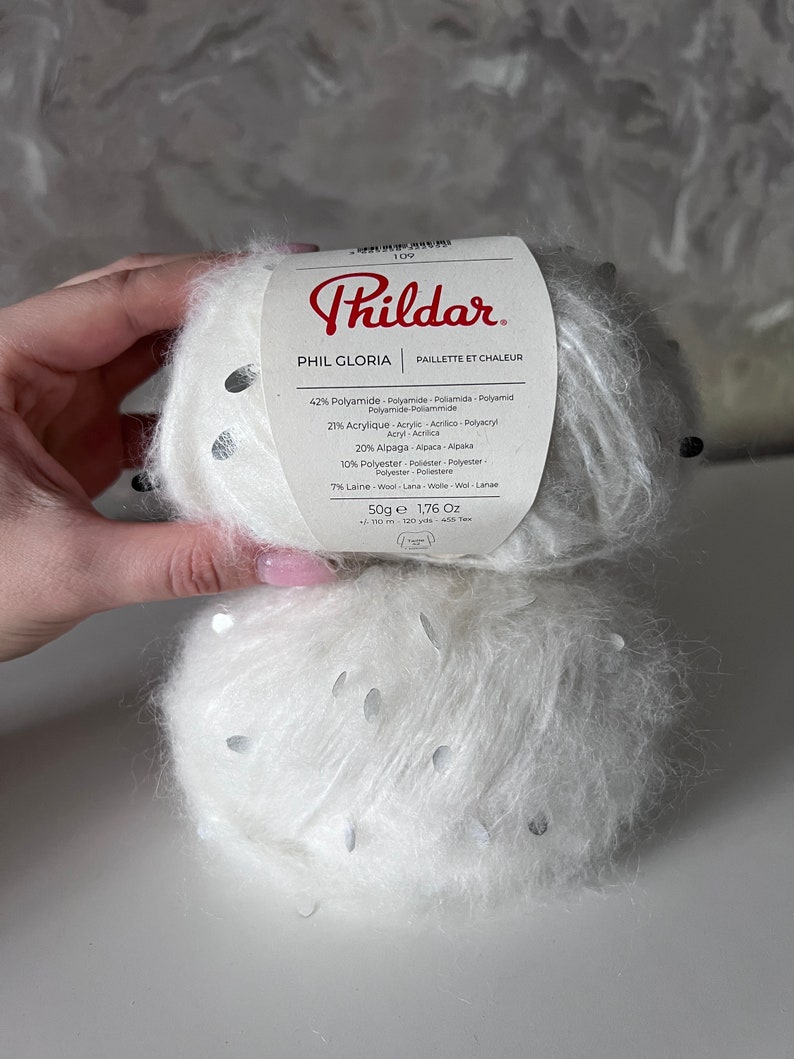 Pu&ograve; includere: Un gomitolo di filato bianco con paillettes argentate, etichettato "Phil Gloria" con una descrizione della composizione del filato: 42% Poliammide, 27% Acrilico, 20% Alpaca, 10% Poliestere, 7% Lana. L'etichetta riporta anche "Paillete et Chaleur" e "50g e 1,76 Oz".