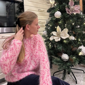 May include: A woman wearing a pink and white fuzzy sweater with a leopard print pattern. She is sitting in front of a decorated Christmas tree.