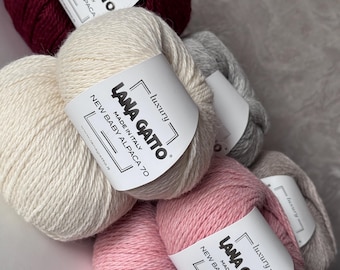 Könnte beinhalten: Vier Knäuel Garn in verschiedenen Farben: Burgunderrot, Creme, Grau und Rosa. Jedes Knäuel hat ein weißes Etikett mit dem Text "LANA GATTO" und "NEW BABY ALPACA 70".