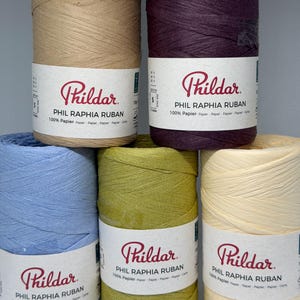 May include: Five spools of Pildar Phil Raphia Ruban 100% paper yarn in tan, dark purple, light blue, olive green, and cream. Each spool features a white label with the brand name Pildar, the product name, and the material composition in multiple languages.