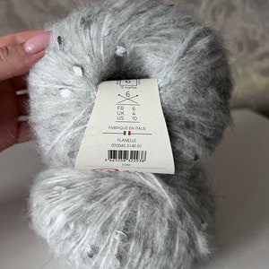 Pu&ograve; includere: Un gomitolo di filato grigio con macchie argentate. L'etichetta del filato riporta "Flanelle 070045 2140 01" e include raccomandazioni per la dimensione degli aghi in misure francesi, britanniche e statunitensi.