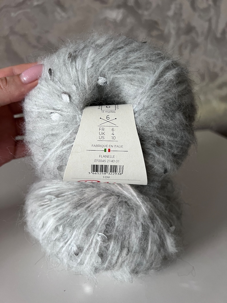 Pu&ograve; includere: Un gomitolo di filato grigio con macchie argentate. L'etichetta del filato riporta "Flanelle 070045 2140 01" e include raccomandazioni per la dimensione degli aghi in misure francesi, britanniche e statunitensi.