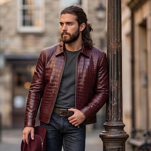 May include: A deep burgundy leather jacket with a textured crocodile-skin pattern, featuring a classic collar and front zipper. The jacket is worn over a dark gray t-shirt and dark blue denim jeans with a brown leather belt, holding a matching hat.