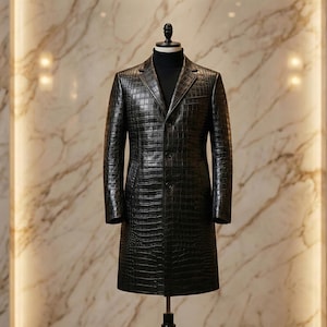 May include: A black, long leather coat with a crocodile skin texture is displayed on a mannequin. The coat has a classic design with a notched collar and button closure. The mannequin is on a black stand in front of a marble wall.
