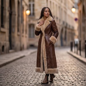 May include: A long, brown, crocodile-textured leather coat featuring a thick, cream-coloured faux fur trim along the lapels, cuffs, and hem. The coat is worn with brown leather ankle boots, creating a structured, full-length outer garment.