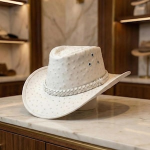 Pode incluir: Um chapéu de cowboy branco com uma superfície texturizada e uma faixa trançada. O chapéu tem uma aba larga e uma coroa estruturada com três pequenos orifícios de ventilação. O chapéu é exibido em uma superfície de mármore.