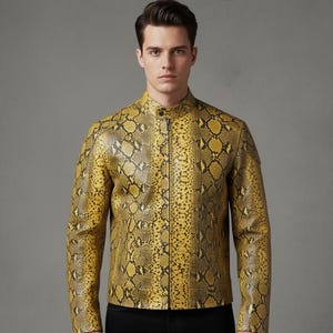 Könnte beinhalten: Gelb-schwarze Jacke mit Schlangenmuster, Stehkragen und Frontreißverschluss. Die Jacke wird über einer schwarzen Hose getragen. Das Model steht vor einem grauen Hintergrund.
