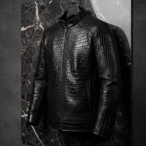 Puede incluir: Chaqueta de cuero negro con textura de piel de cocodrilo. La chaqueta tiene cuello alto, cremallera frontal y un bolsillo. Cuelga de una percha negra sobre un fondo oscuro.