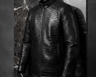 May include: A black leather jacket with a crocodile skin texture. The jacket has a stand-up collar, a front zipper, and a single pocket. The jacket is hanging on a black hanger against a dark background.