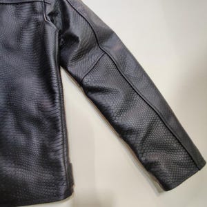 Może przedstawiać: Czarna kurtka ze sztucznej sk&oacute;ry o fakturze sk&oacute;ry węża. Kurtka ma klasyczny kr&oacute;j z dopasowanym krojem, widocznymi szwami i strukturalną sylwetką. Rękaw jest ustawiony pod kątem, prezentując szczeg&oacute;łowy wz&oacute;r.