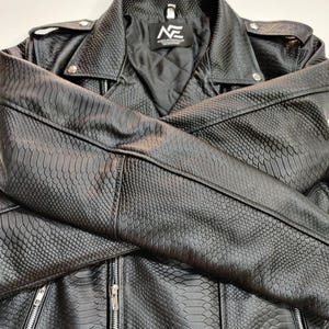 Może przedstawiać: Czarna kurtka ze sztucznej sk&oacute;ry o fakturze sk&oacute;ry węża. Kurtka posiada kołnierz z klapami, srebrne zamki błyskawiczne i pikowaną podszewkę. Logo marki jest widoczne na metce wewnątrz kurtki.