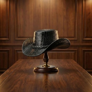 May include: A black cowboy hat with a crocodile-effect texture is displayed on a wooden stand with a brass base. The hat features a braided band and a wide brim. The hat is on a dark wooden table.