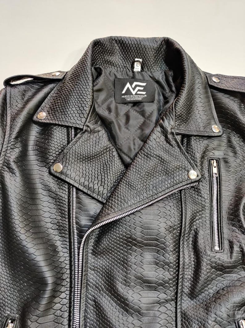 Może przedstawiać: Czarna kurtka motocyklowa ze sztucznej sk&oacute;ry o fakturze sk&oacute;ry węża. Kurtka posiada kołnierz z klapami, srebrne zamki błyskawiczne i zatrzaski. Wewnętrzna podszewka jest czarna i pikowana. Widoczne jest logo marki "NE".