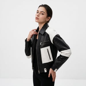 Pode incluir: Um casaco de couro preto e branco com gola clássica, fecho de correr frontal completo e vários bolsos com fecho. O design inclui painéis de couro branco nos ombros e um bolso branco grande no lado esquerdo da parte frontal.