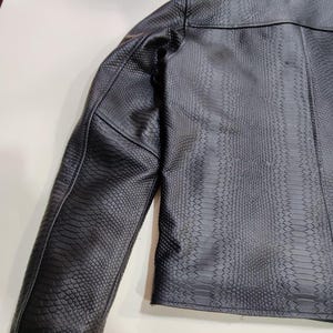 Może przedstawiać: Czarna kurtka ze sztucznej sk&oacute;ry o fakturze sk&oacute;ry węża. Kurtka ma klasyczny kr&oacute;j z wykładanym kołnierzem i dopasowanym krojem. Rękawy są długie, a og&oacute;lny styl jest odważny i nowoczesny.