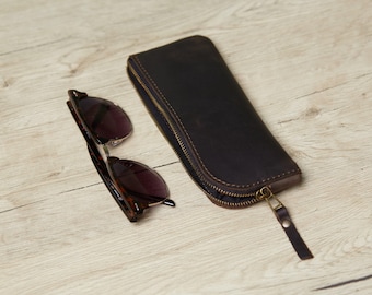 May include: A dark brown leather zippered case with visible stitching and a matching leather pull tab, positioned next to a pair of dark-tinted sunglasses with a tortoiseshell-patterned frame, both items resting on a light-colored wood grain surface.