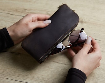 May include: A dark brown leather zippered case with visible stitching along the edges, holding a pair of sunglasses with dark lenses and a tortoiseshell frame. The case has a rounded top edge and a metal zipper closure along the side.