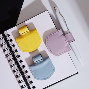 May include: Three small, semi-circular pen holders in yellow, lavender, and light blue, each featuring a stitched loop for a pen. A white pen is inserted into the lavender holder, and all three are positioned on a white spiral-bound notebook.