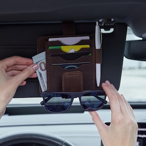 Könnte beinhalten: Ein dunkelbrauner Auto-Sonnenblenden-Organizer aus Leder mit mehreren Fächern für Kreditkarten, einer seitlichen Schlaufe für einen weißen Stift, einem vertikalen Fach mit einem Hundert-Dollar-Schein und einem unteren Riemen für eine schwarze Sonnenbrille.