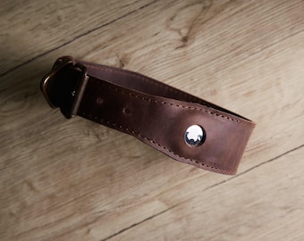 May include: A dark brown leather collar featuring a metallic buckle and a circular silver-toned hardware accent embedded in the center. The collar is constructed with visible stitching along the edges and includes multiple adjustment holes.