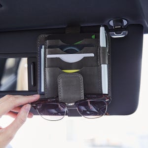 May include: A dark grey leather car visor organiser featuring multiple vertical slots for credit cards or identification, a dedicated side loop for a pen, and a central strap designed to hold a pair of dark-rimmed eyeglasses securely in place.