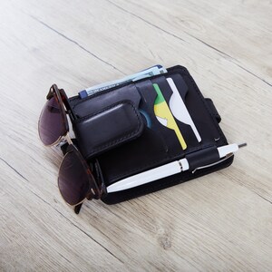 May include: A black leather wallet featuring multiple card slots, a magnetic money clip, and a pen loop. The wallet holds a white pen, several credit cards, and currency. A pair of tortoiseshell sunglasses is attached to the front exterior.