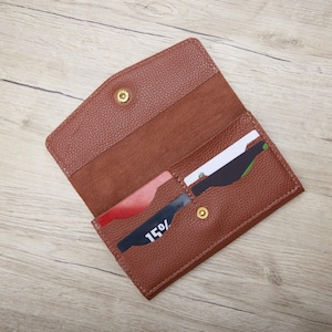 May include: A brown leather wallet, open to show card slots and a press stud closure. The wallet has a textured finish and contains several cards, including one with "15%" printed on it. The wallet is on a wooden surface.