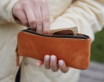 May include: A rectangular tan leather zippered pouch with visible stitching along the edges. The pouch features a black interior lining and a metal zipper closure. A pair of light-colored eyeglasses is partially visible inside the open pouch.