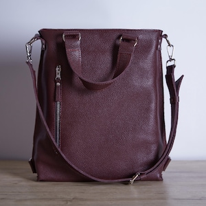 Puede incluir: Una mochila de cuero texturizado en color burdeos intenso con dos asas superiores, un bolsillo vertical con cremallera en la parte frontal y correas ajustables para los hombros sujetas con clips de metal plateado a los lados.
