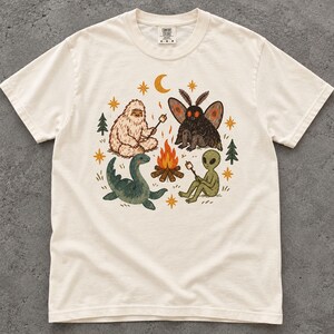 Può includere: Una t-shirt a maniche corte color bianco sporco con una stampa grafica raffigurante un Bigfoot, un uomo falena, un mostro di Loch Ness e un alieno seduti attorno a un falò, con piccoli pini, stelle e una luna crescente nei toni dell'arancio e del verde.